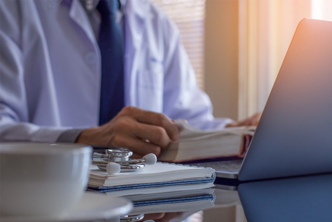 A person in a white coat sits at a desk with a laptop, notebook, stethoscope, and book, diligently focused on AMC Exam Preparation. A cup is visible in the foreground, hinting at long study hours fueled by coffee and determination.