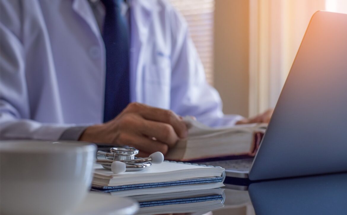 A person in a white coat sits at a desk with a laptop, notebook, stethoscope, and book, diligently focused on AMC Exam Preparation. A cup is visible in the foreground, hinting at long study hours fueled by coffee and determination.