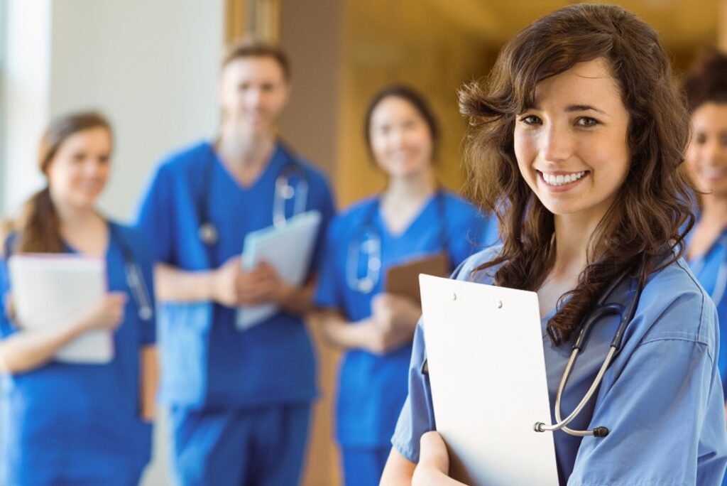 A group of medical professionals in blue scrubs stands together, with one person holding a clipboard in the foreground, discussing changes to the MCCQE Part I in 2025 and referencing Ace QBank to align with new MCC objectives.