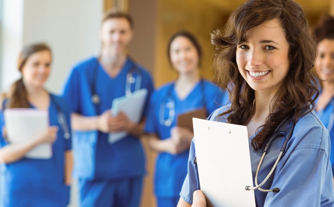 A group of medical professionals in blue scrubs stands together, with one person holding a clipboard in the foreground, discussing changes to the MCCQE Part I in 2025 and referencing Ace QBank to align with new MCC objectives.