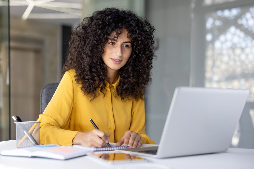 A person with curly hair, wearing a yellow shirt, writes in a notebook while exploring Ace QBank on their laptop in an office setting preparing for the MCCQE1 exam.