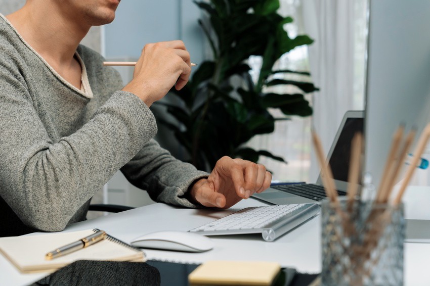 A person in a gray sweater sits at a desk, diligently preparing for the MCCQE1 exam. They're holding a pencil while using the keyboard, likely accessing Ace QBank's comprehensive question bank. Nearby are a notebook, pen, and container with pencils; a plant adds warmth to the background.