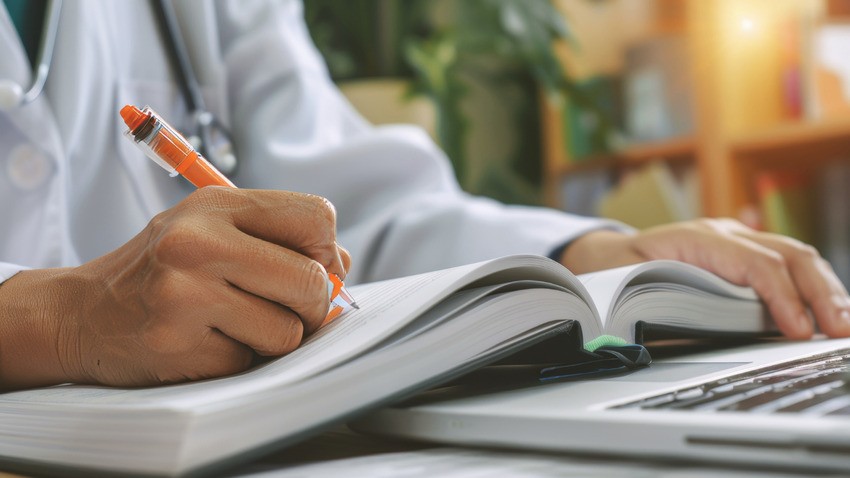 A person in a lab coat writes in an open book while using a laptop, perhaps strategizing for the AMC Exam Preparation, with blurred background elements.