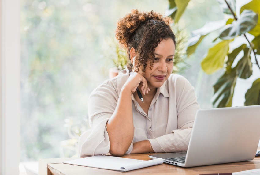 A woman with curly hair sits at a wooden table, focused on studying for the MCCQE1 exam using Ace QBank. A notebook and pen are nearby, ready to capture insights. Large green plant leaves create a serene background as she immerses herself in her preparations.