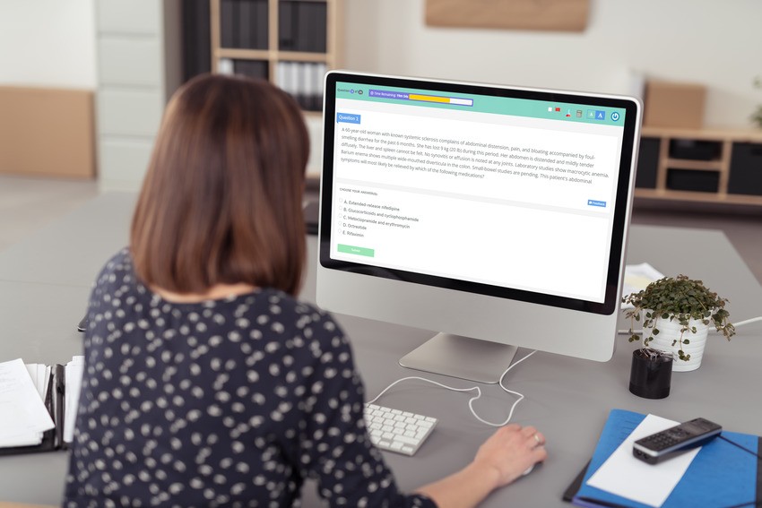 A person sits at a desk, engrossed in AMC Exam Preparation. The desktop screen glows with a text document outlining effective study techniques for the AMC exam in bullet points. An array of office items and a question bank are neatly arranged on the desk, ready to assist in their focused study session.