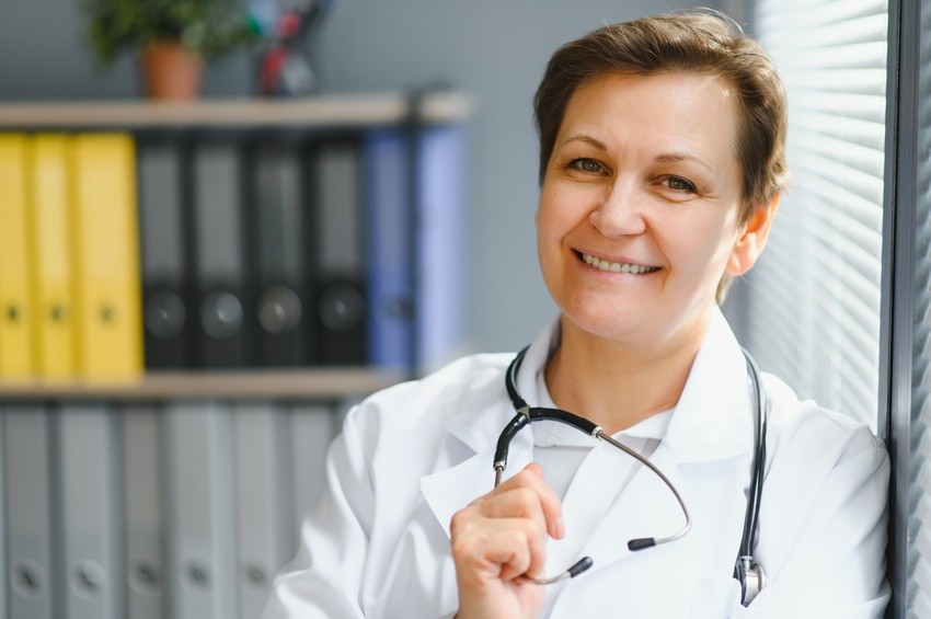 A smiling person in a white coat with a stethoscope around their neck stands in an office adorned with shelves of colorful binders, perhaps contemplating the Ace QBank—renowned as the best Canadian question bank for MCCQE1 exam preparation.