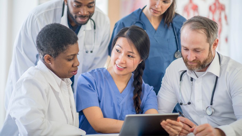 A group of medical professionals, including doctors and nurses, gathered around a tablet device and engaged in a discussion about the upcoming changes to the MCCQE Part I in 2025.