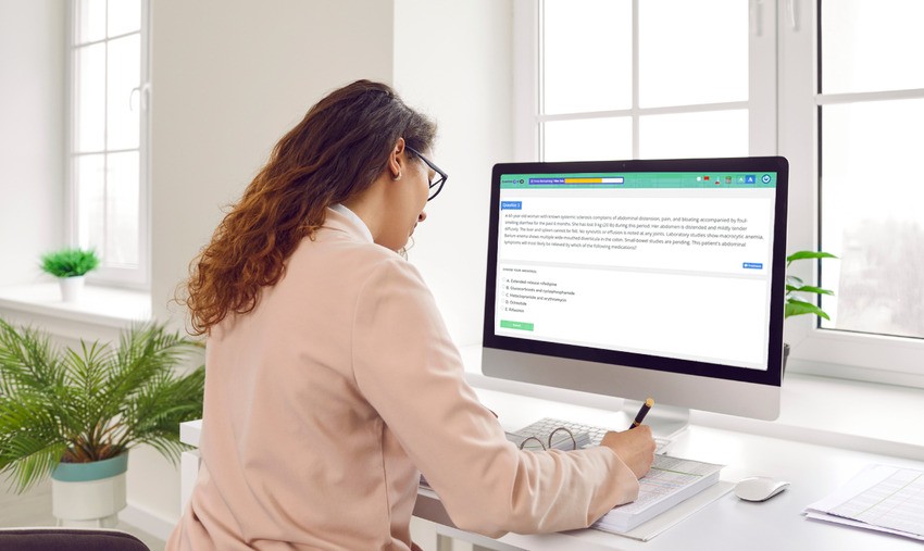 A person in a white shirt is sitting at a desk, taking notes while preparing for the MCCQE1, focused on information displayed on their computer screen. A potted plant adds a touch of nature to the desk, and large windows reveal an inspiring view in the background.