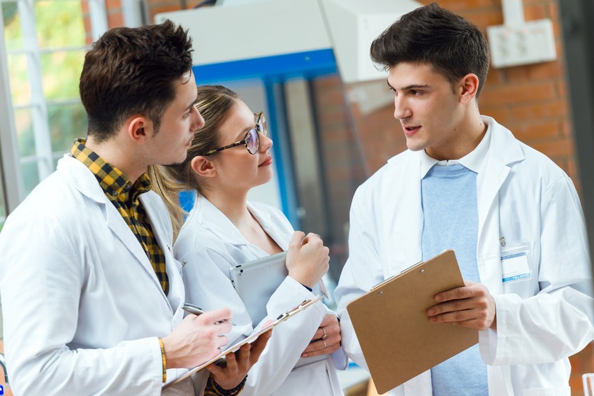 In a laboratory setting, three individuals in lab coats discuss the MCCQE1 exam dates while holding clipboards. They're deeply engaged, possibly reviewing strategies from Ace QBank to ensure success.