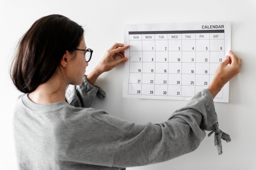 A person with glasses hangs a calendar on a white wall, eagerly circling MCCQE1 exam dates to begin their focused study sessions with Ace QBank.