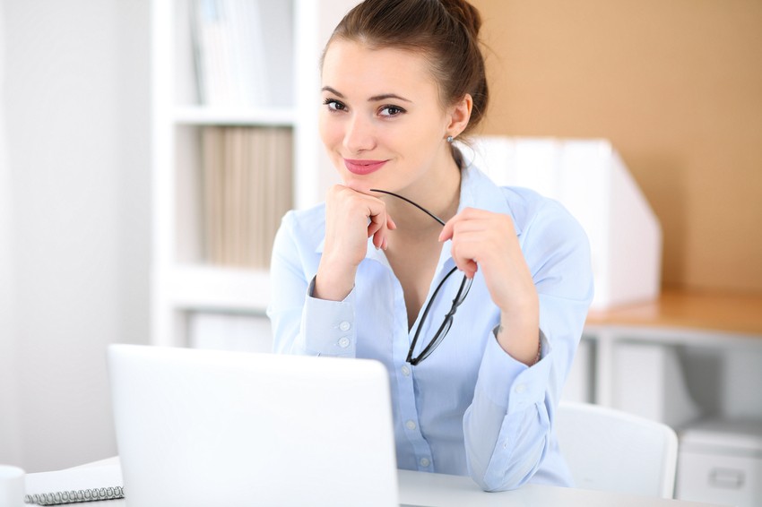 A smiling woman in a blue shirt sits at a desk, holding glasses, with a laptop in front of her—perhaps she's exploring the best Canadian question bank for MCCQE1 or uncovering MCCQE1 Exam Passing Secrets for 2025.