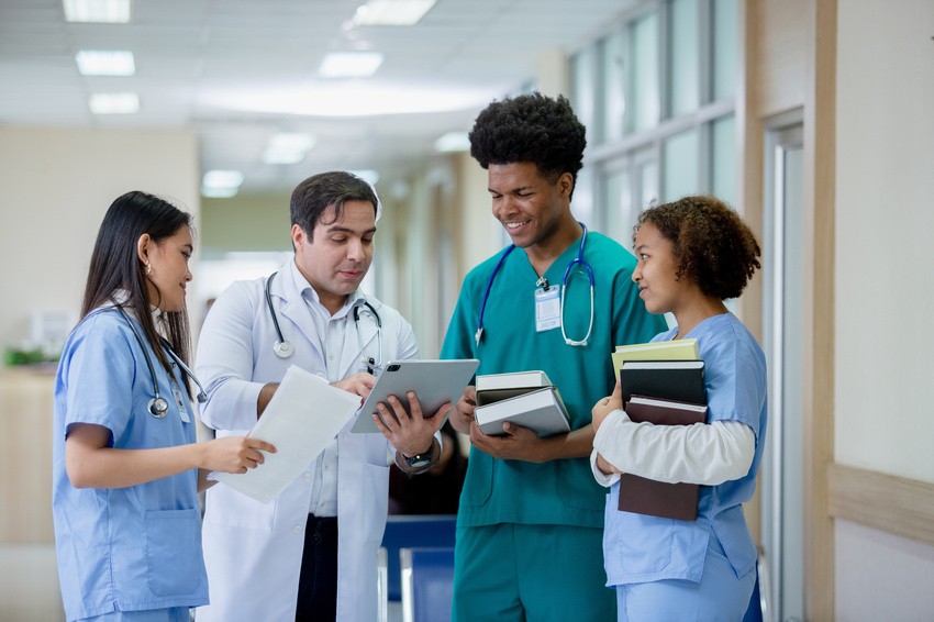 A group of medical professionals in a hospital corridor looks at a tablet, preparing for the MCCQE1 exam. Two hold books while another holds papers, all focused and engaged in discussing the MCC Objectives critical for MCCQE1 2025 success.