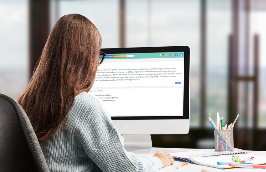 A woman with long brown hair sits at a desk using a desktop computer, following a step-by-step guide for IMGs. A window with a blurred outdoor view is in the background, and pencils and an Ace QBank notebook are on the desk.