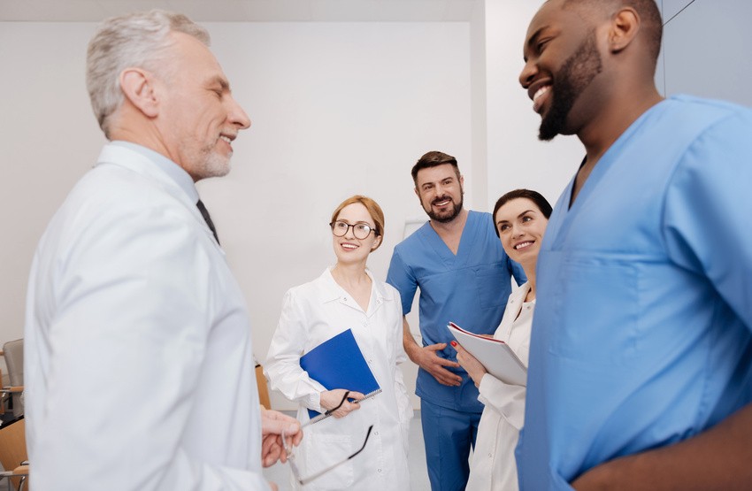 A group of healthcare professionals in white coats and blue scrubs are standing together, smiling and holding documents in a clinical setting. Perhaps they're celebrating after acing the MCCQE1 2025 using Ace QBank's step-by-step guide for IMGs.