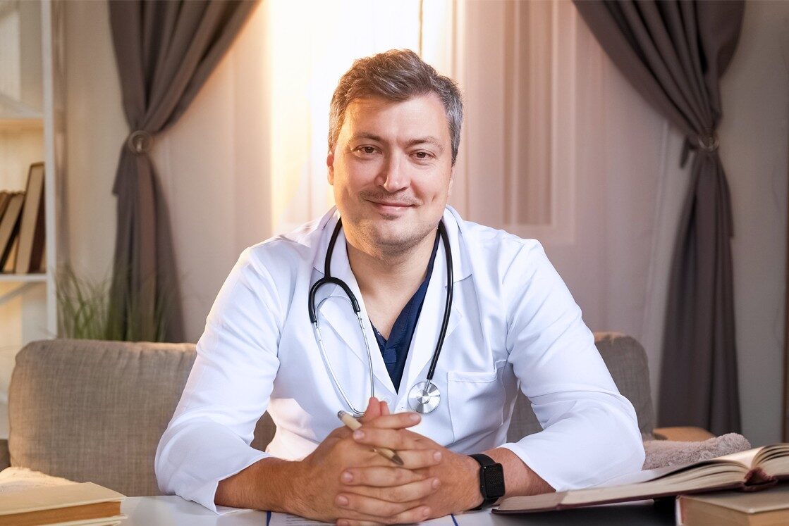 A doctor in a white coat sits at a desk with a stethoscope around his neck, ready to help you crush the 2025 MCCQE1 on your first try. His confident smile suggests he's armed with knowledge from Ace QBank, surrounded by books and curtains.