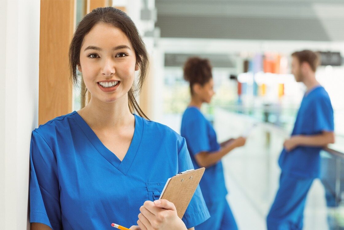 A woman in blue scrubs holds a clipboard, smiling confidently, perhaps gearing up for MCCQE1 2025. In the background, two colleagues in similar attire discuss Ace QBank, their chosen tool for MCCQE1 exam preparation.