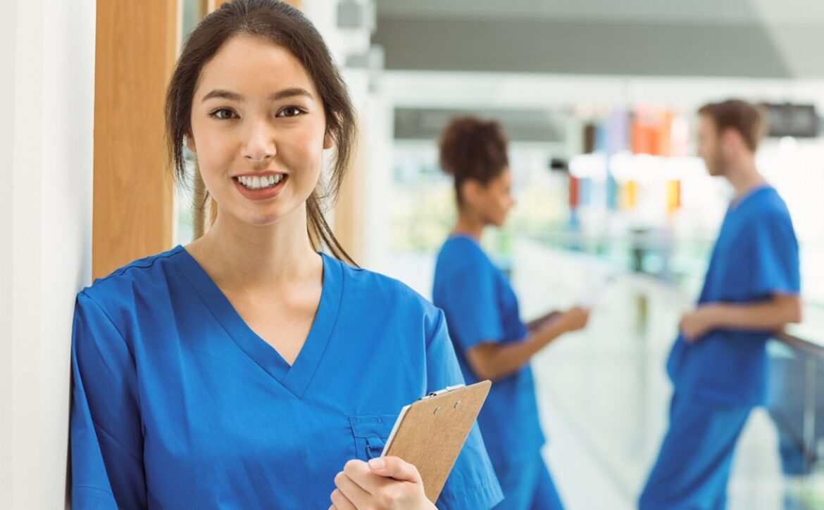 A woman in blue scrubs holds a clipboard, smiling confidently, perhaps gearing up for MCCQE1 2025. In the background, two colleagues in similar attire discuss Ace QBank, their chosen tool for MCCQE1 exam preparation.