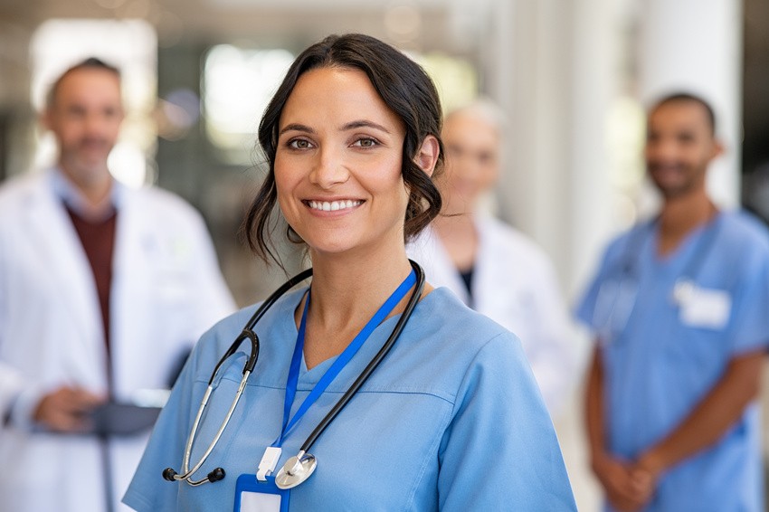 A smiling nurse in blue scrubs with a stethoscope around her neck stands in the foreground, embodying the dedication required to succeed in exams like MCCQE1 2025, while three medical professionals work diligently in the blurred background.