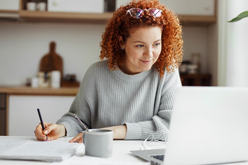 A person with curly red hair, wearing a gray sweater, writes diligently in a notebook while looking at a laptop in a bright kitchen, diving deep into their MCCQE1 exam preparation.