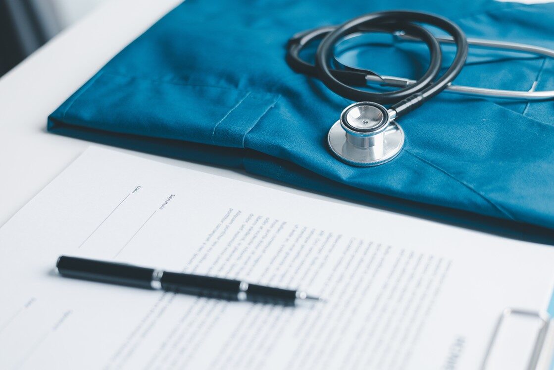 A stethoscope rests on blue medical scrubs beside Ace QBank materials. A pen lies on a document about the Step-by-Step Guide for IMGs, creating a workspace with medical and administrative items perfect for preparing for MCCQE1 2025 on the white table.