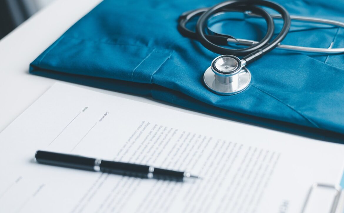 A stethoscope rests on blue medical scrubs beside Ace QBank materials. A pen lies on a document about the Step-by-Step Guide for IMGs, creating a workspace with medical and administrative items perfect for preparing for MCCQE1 2025 on the white table.