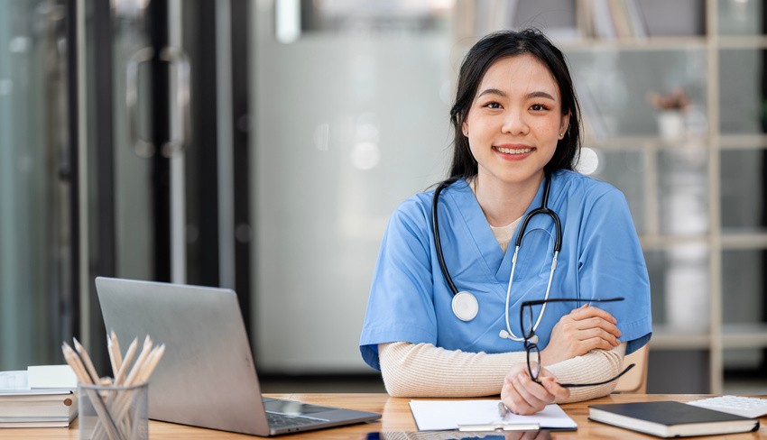 A person in blue scrubs sits at a desk with a laptop and documents, pondering "How Can I Score High in MCCQE1 2025?" while holding glasses and smiling with confidence.