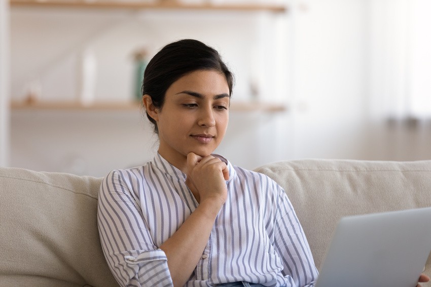 A woman in a striped shirt sits on a couch, immersing herself in Ace QBank on her laptop with a thoughtful expression, preparing for the AMC MCQ exam. Shelves are neatly arranged in the background.