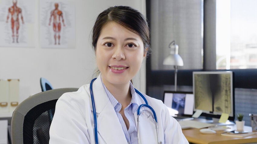 A person in a white coat with a stethoscope, sitting in an office with anatomical charts and a computer displaying the Step-by-Step Guide for IMGs preparing for the MCCQE1 2025.