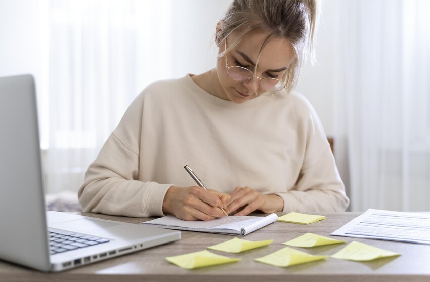 A person wearing glasses is engrossed in MCCQE1 2025 exam preparation, diligently writing in a notebook at a desk cluttered with sticky notes, a laptop, and papers. Ace QBank materials are scattered around, reflecting their dedication to acing the upcoming challenges.